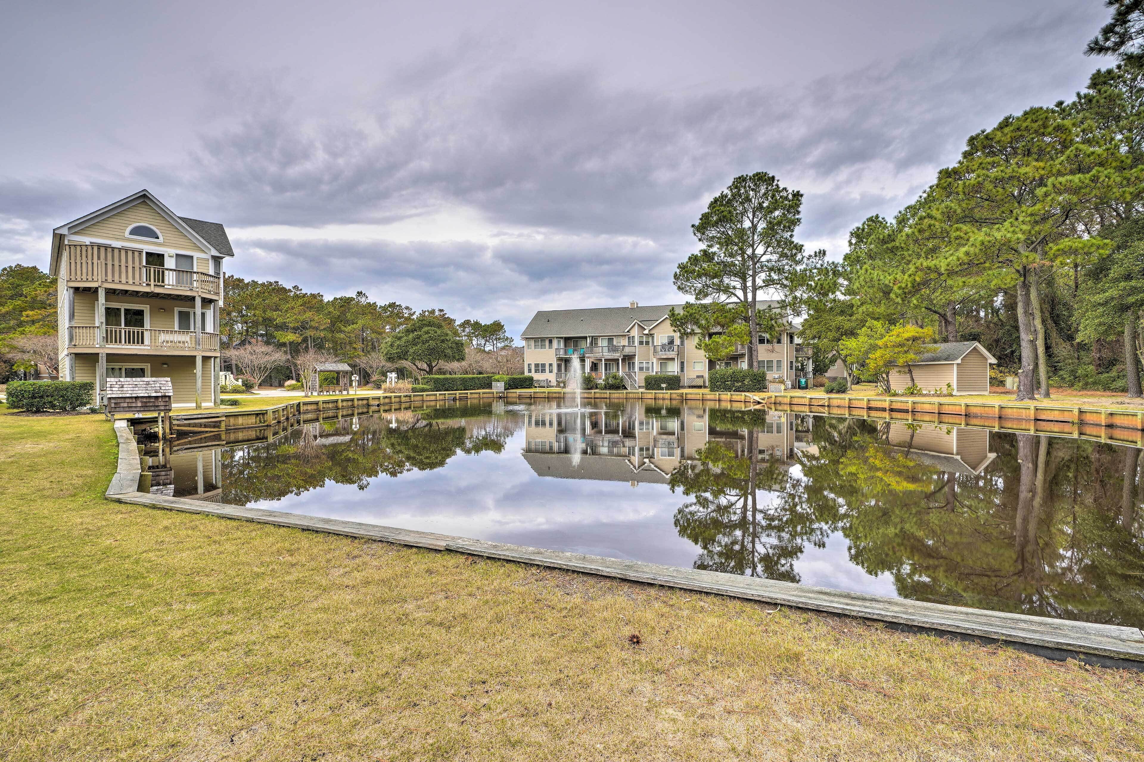 A multi-story building with a balcony, a pond with a wooden bridge, and surrounding trees.