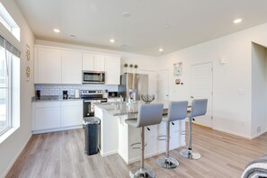 A modern kitchen with white cabinets, a central island with bar stools, stainless steel appliances, and a large window providing natural light. - Colorado Retreat Near Hiking, Golfing & Fishing! (Commerce City)