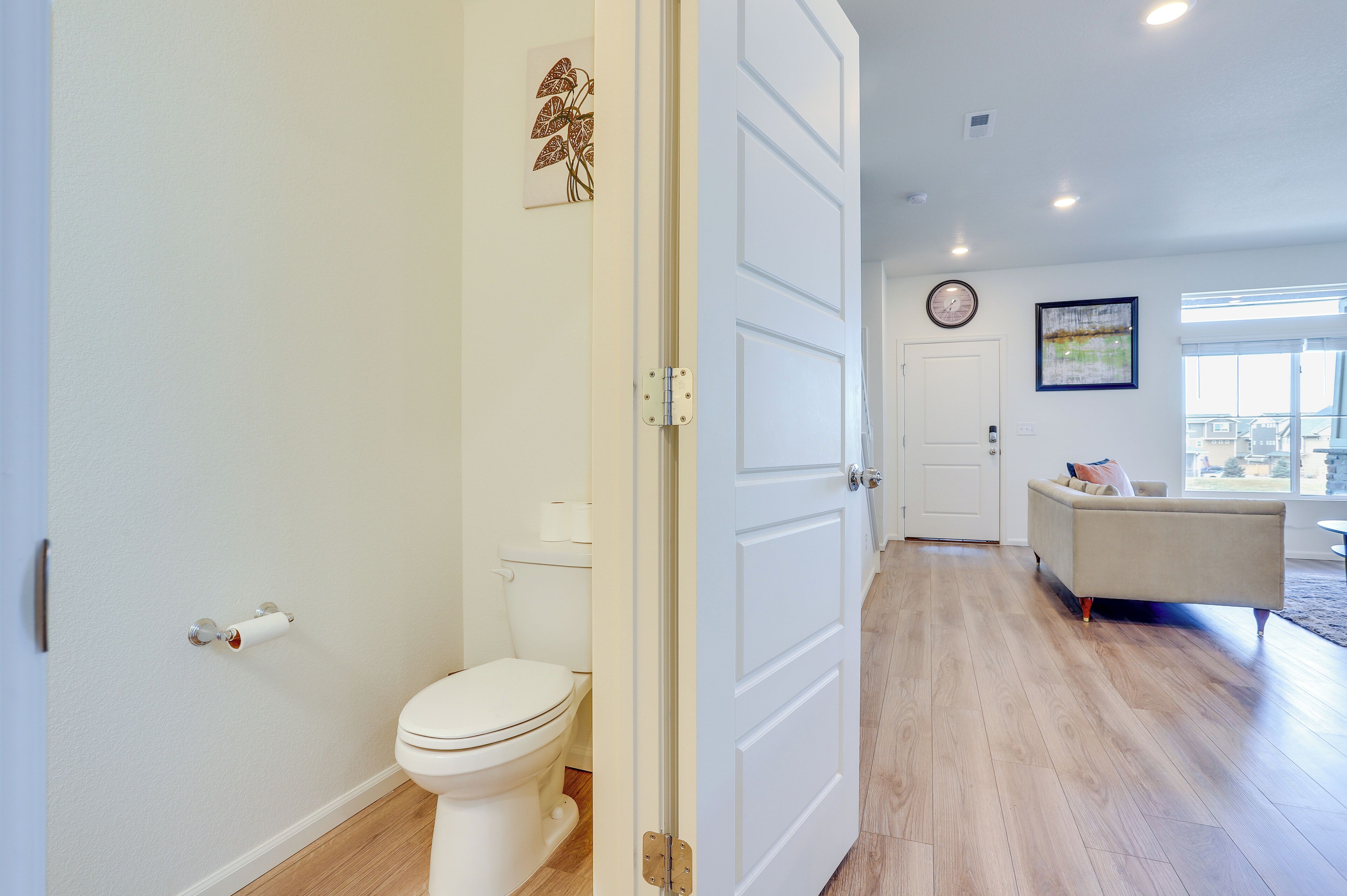 Une salle de bain moderne avec des toilettes, une porte blanche et un couloir menant à un espace de vie comprenant un canapé et une grande fenêtre.