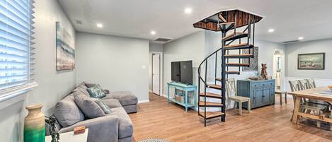 A modern living room with a grey sectional sofa, a round ottoman, a wooden staircase, and a dining area with a table and chairs.