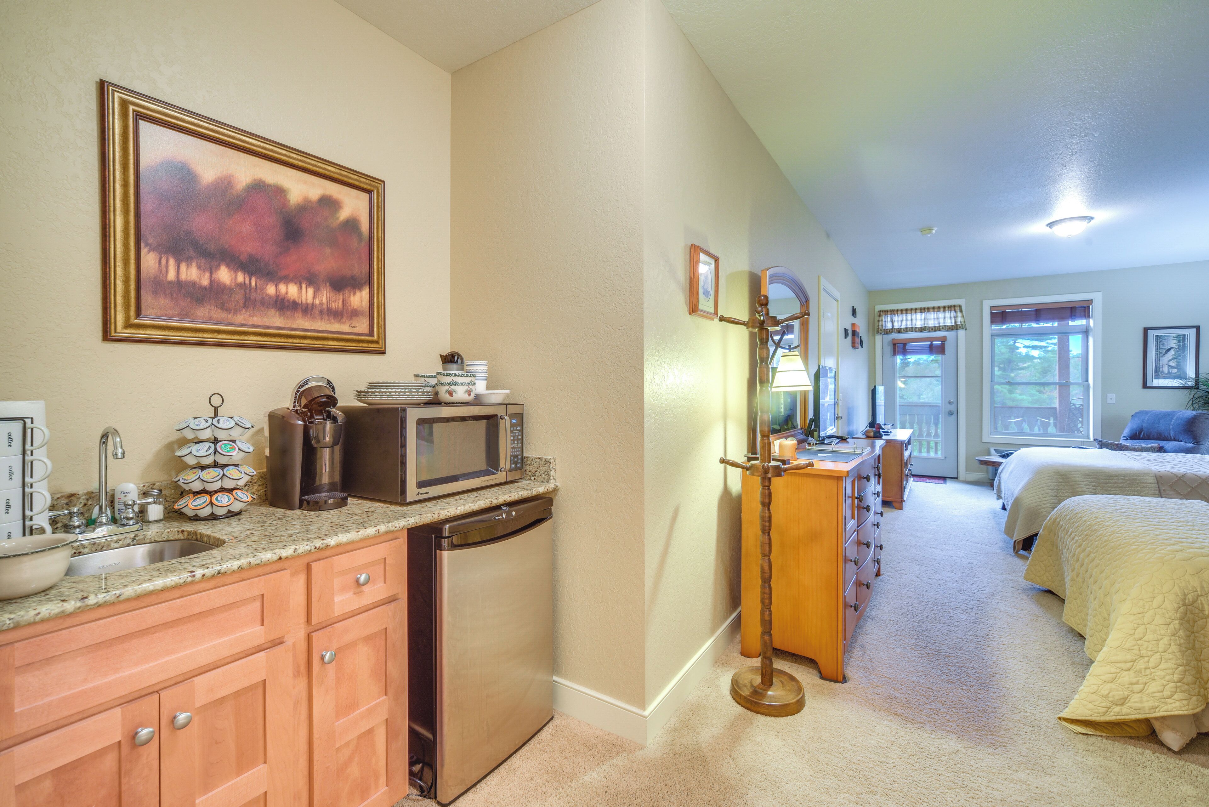 A kitchen with a microwave, sink, and cabinets, a hallway with a dresser and a lamp, and a bedroom with a bed and a window.