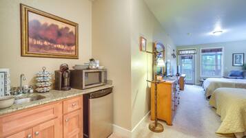 A kitchen with a microwave, sink, and cabinets, a hallway with a dresser and a lamp, and a bedroom with a bed and a window.