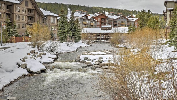 Vista a la orilla del río de una cabaña con varios edificios, una terraza de madera y terreno cubierto de nieve.