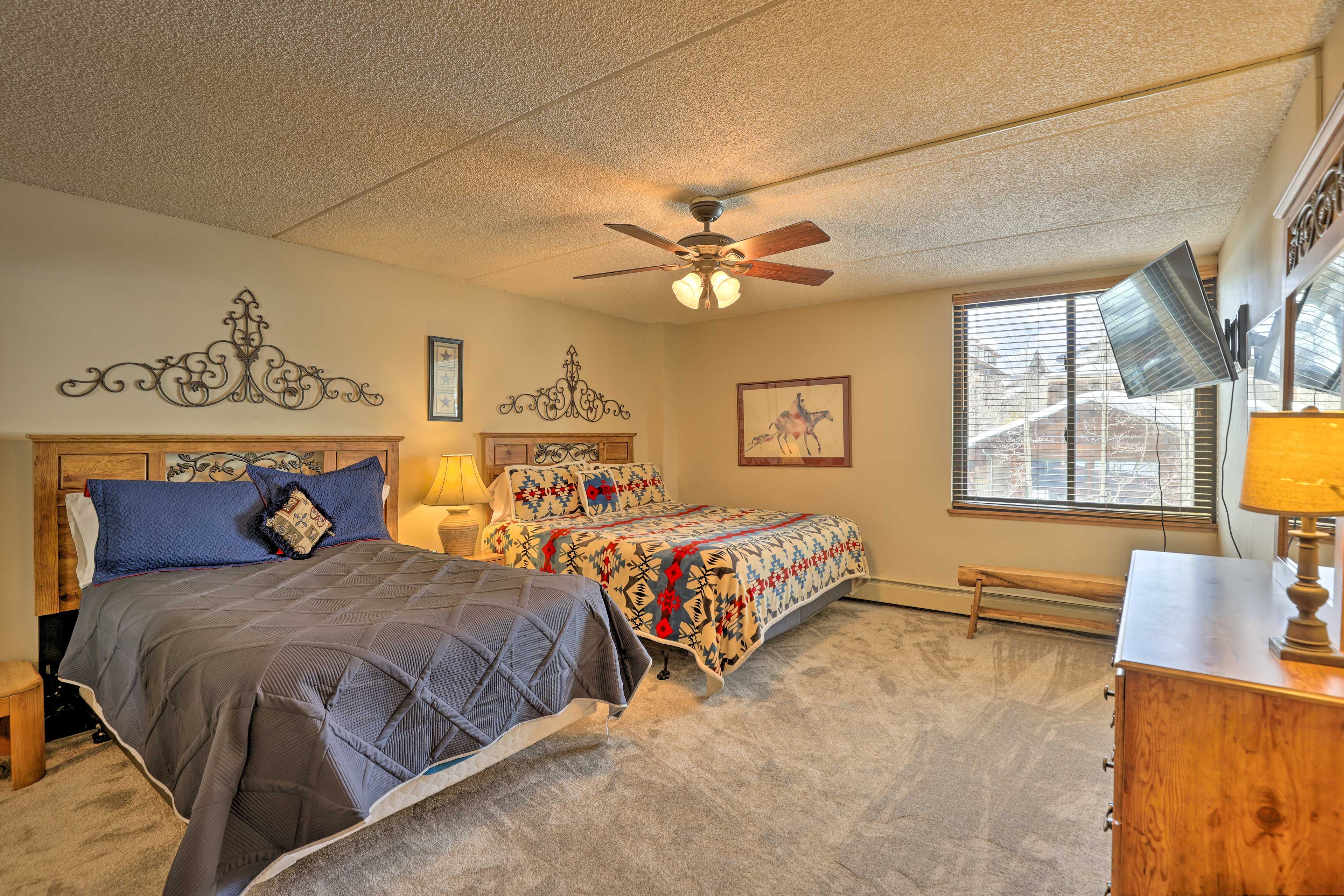 A bedroom with two beds, a ceiling fan, a window with blinds, and a desk with a lamp.