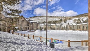 Un espacio exterior cubierto de nieve con una valla de madera, un banco y un edificio de varios pisos al fondo.