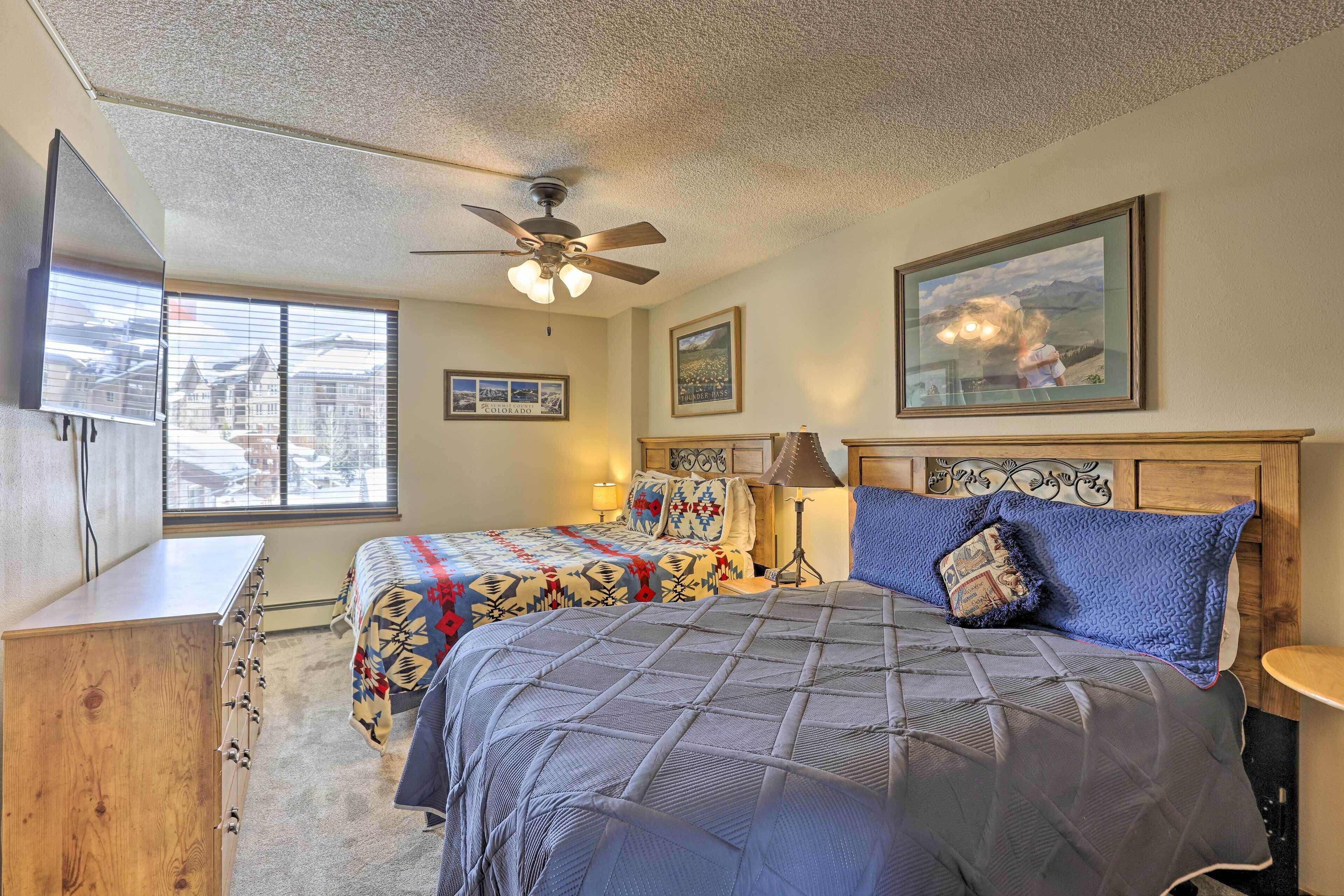 A bedroom with two beds, a ceiling fan, a dresser, and a window with a view of houses.