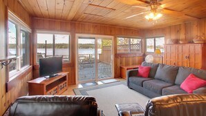 Un acogedor interior de cabaña con techo de madera, un televisor sobre un mueble de madera, un sofá gris con cojines rojos y un amplio ventanal con vista a una terraza.
