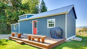 A small, single-story cabin with a red door, a covered picnic table, and a barbecue grill on the deck. - Contemporary Cabin w/ Mountain Views & Deck (Bonners Ferry)