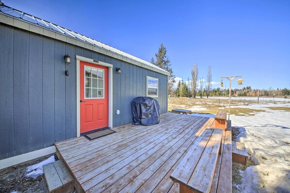 A wooden deck with picnic tables, a red door, and a covered area. - Contemporary Cabin w/ Mountain Views & Deck (Bonners Ferry)