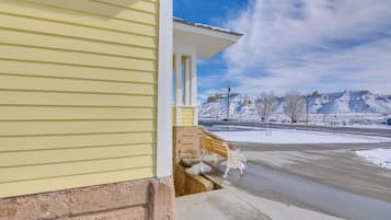 A yellow building with a porch and steps leading to a driveway. Snow-covered mountains and a clear sky are visible in the background.