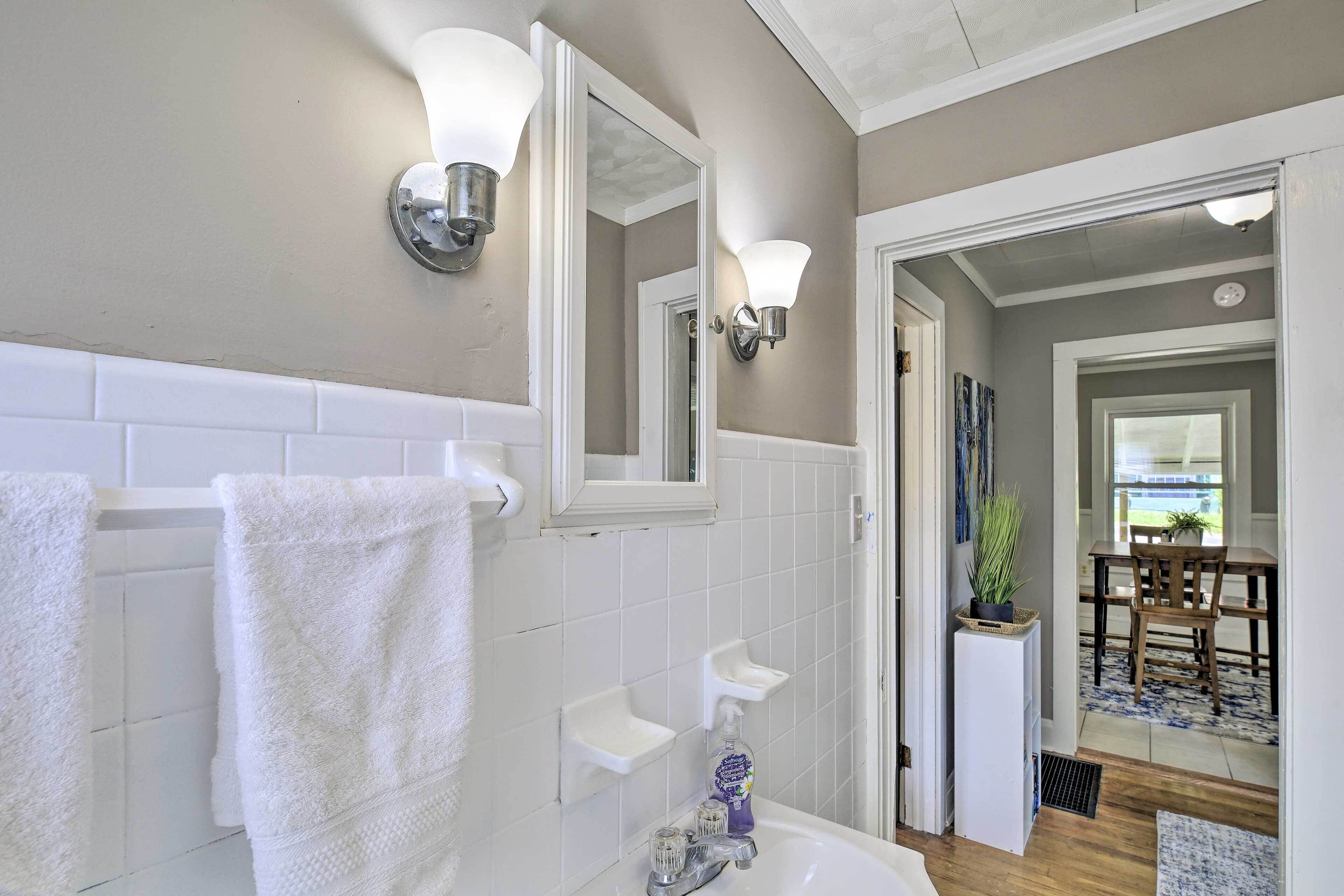 Un baño con azulejos blancos, una bañera blanca, un espejo y un lavabo con dispensador de jabón. Se ve un perchero con toallas blancas y una pequeña planta en un estante.