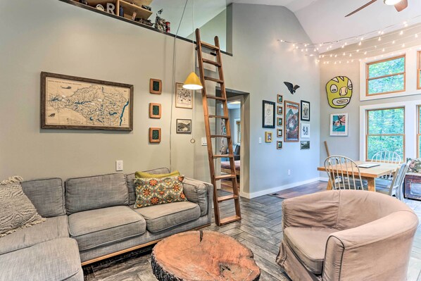 A living room with a gray sofa, a wooden coffee table, a wooden ladder, and a dining area with a wooden table and chairs. - Cozy White Lake Home w/ Patio, Deck & Grill! (White Lake)
