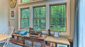 Habitación con amplios ventanales que dan a un bosque, un escritorio de madera con una radio antigua y una silla blanca.