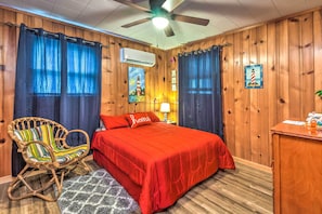 A room with a wooden paneled wall, a bed with a red bedspread, a wicker chair, and a desk with a lamp.