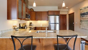 A kitchen with wooden cabinets, stainless steel appliances, granite countertops, and two bar stools. - Alluring Manson Lakeside Condo w/ Shared Pool! (Manson)