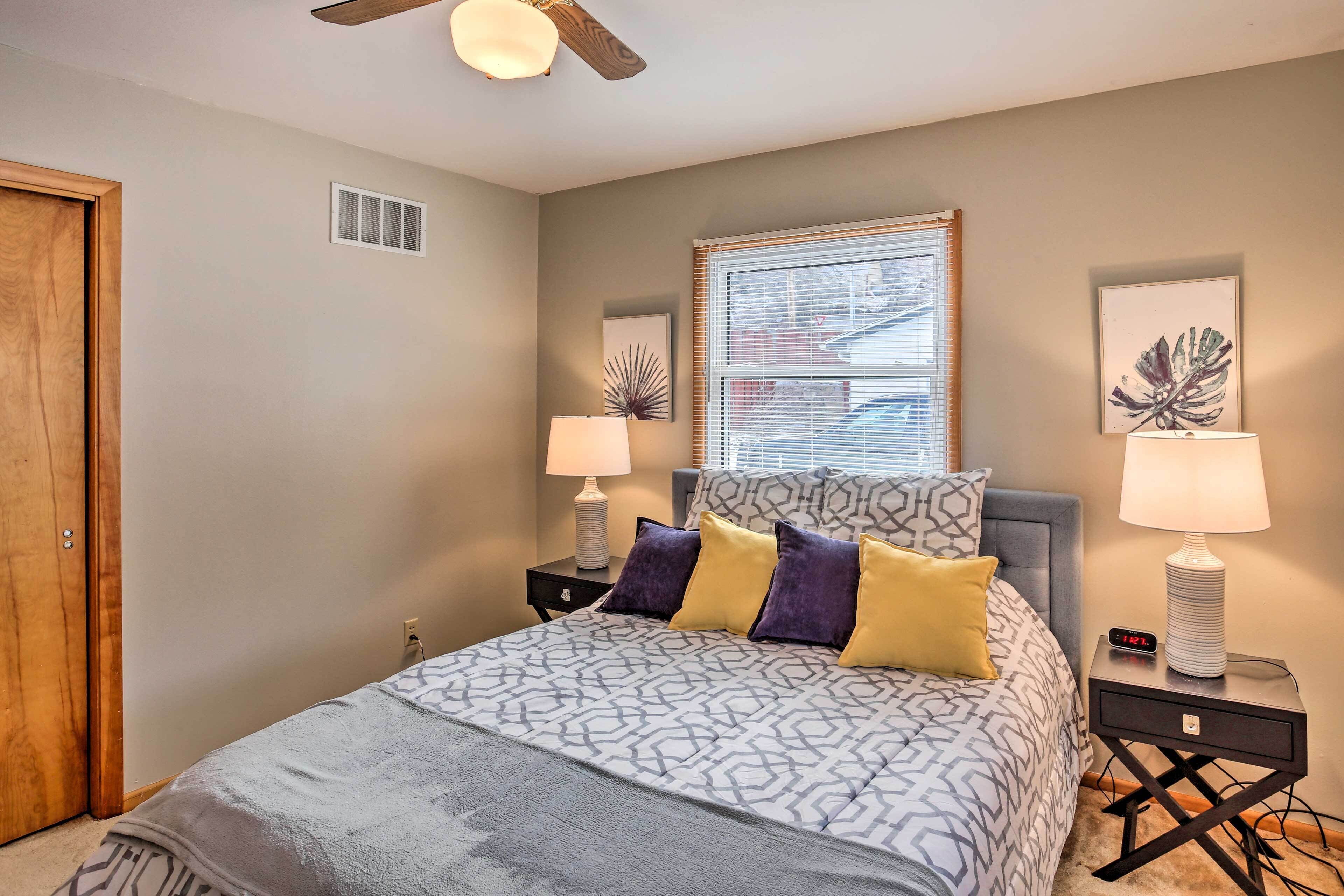 A bedroom with a bed, two bedside tables, a ceiling fan, a window with blinds, and a door.