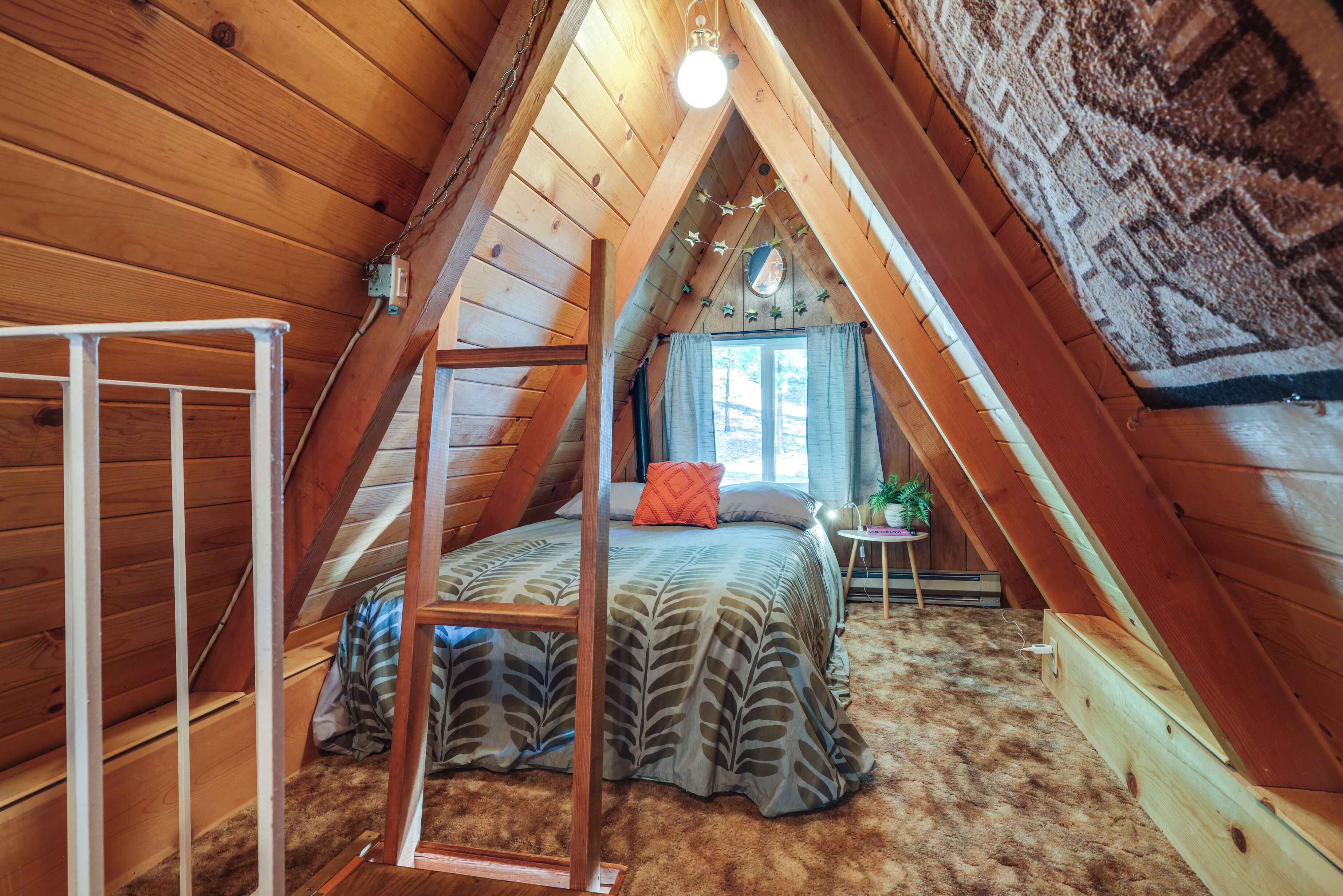 A cozy attic bedroom with a sloped ceiling, a bed with a leaf-patterned bedspread, a small table with a plant, and a window with curtains.