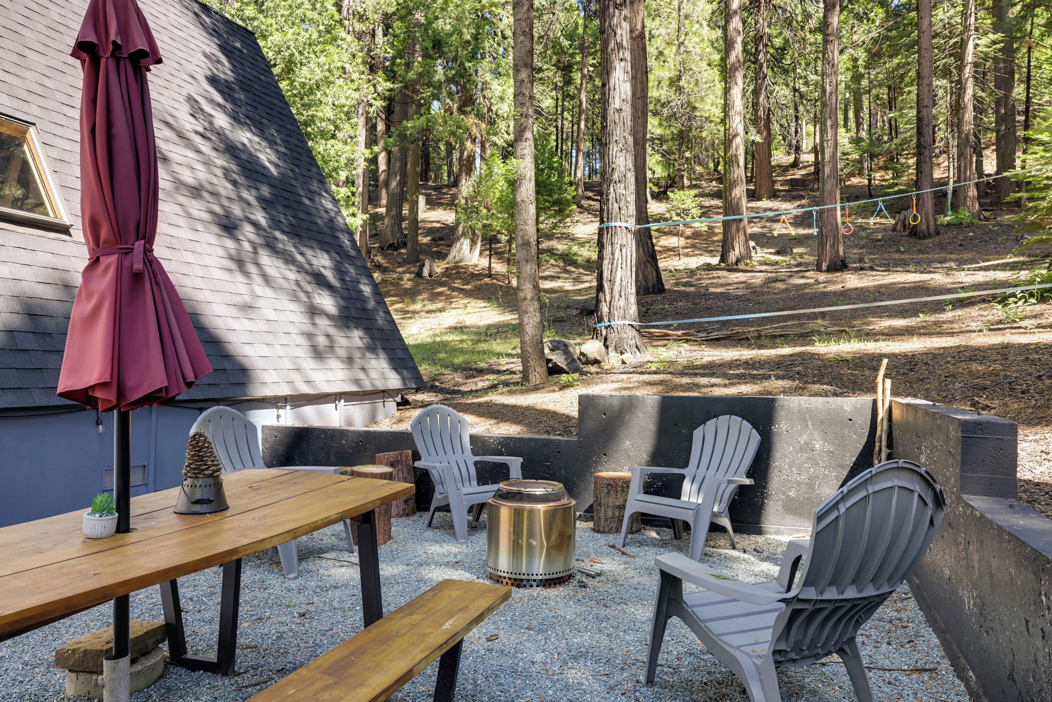 Eine Außenpatio mit einem Holztisch, zwei Adirondack-Stühlen, einer Feuerstelle aus Metall und einem roten Sonnenschirm.