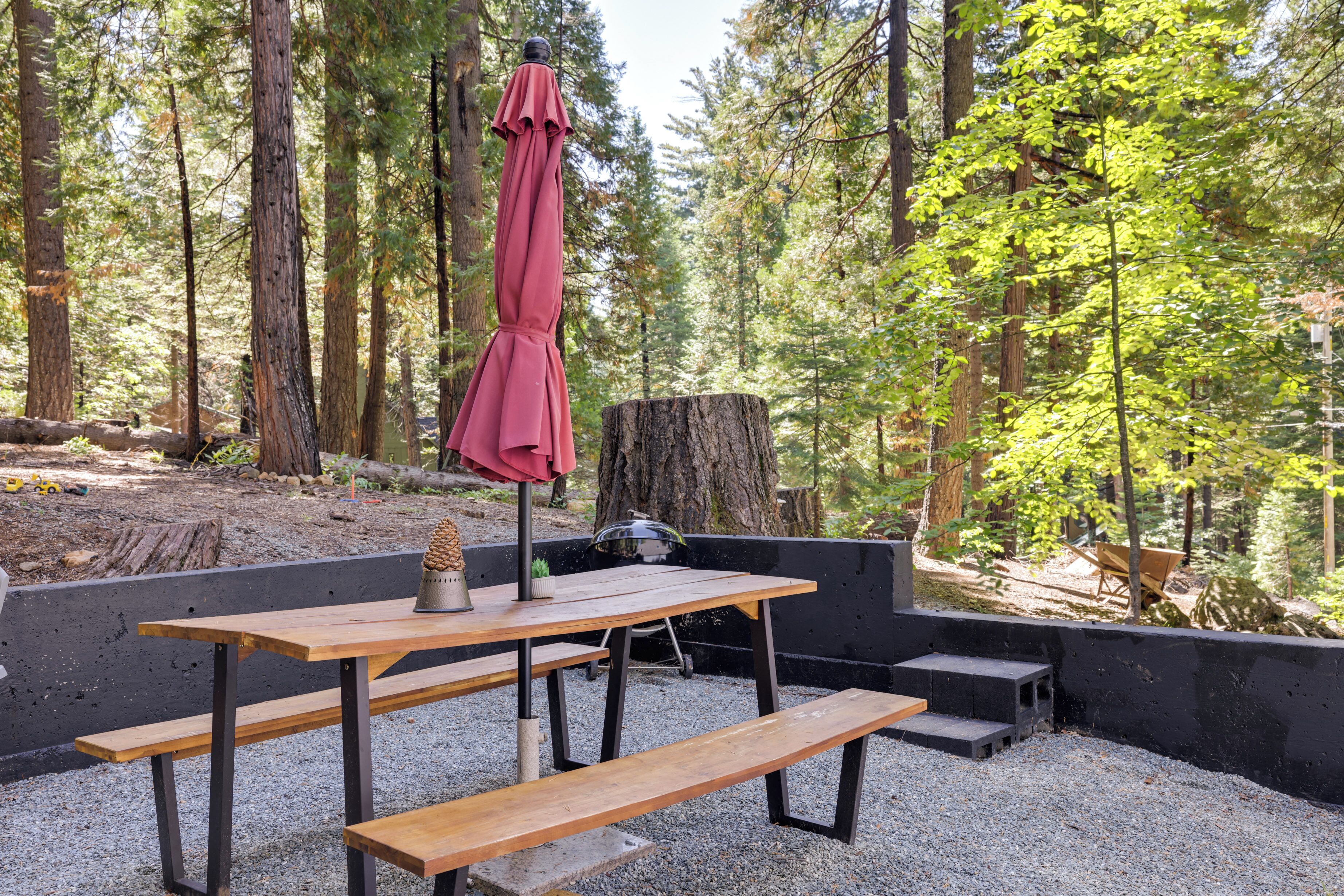 Une table en bois installée à l’extérieur, avec des bancs, un parasol rouge et un barbecue, le tout situé dans une zone boisée.