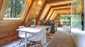 A wooden cabin interior with a dining area, white chairs, a table, and a large window overlooking a forest. - Decks, Grills + Fire Pit: Camp Connell Hideaway! (Arnold)