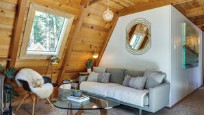 A cozy living room with a glass coffee table, a white sofa, a fluffy chair, and a large window offering a view of trees. - Decks, Grills + Fire Pit: Camp Connell Hideaway! (Arnold)