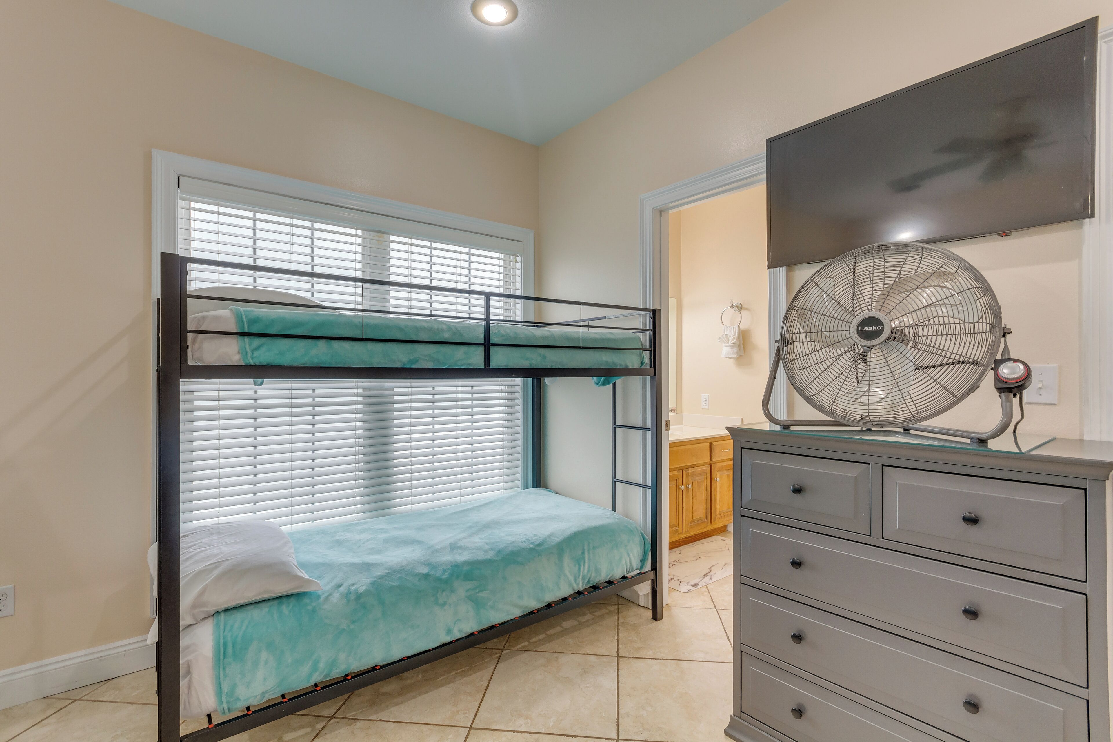 A bunk bed with a fan and a dresser in a room with beige walls and a tiled floor.
