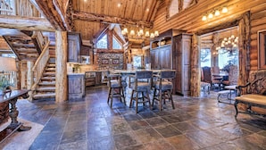 A rustic log cabin interior with a stone-tiled floor, a central island bar, and a staircase leading to an upper level.