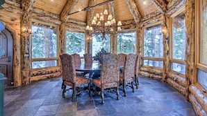 A rustic wooden interior with a large dining table surrounded by ornate chairs, large windows, and a chandelier.