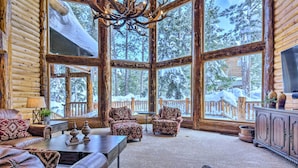 A log cabin interior with large windows, a chandelier, leather furniture, and a view of a snowy landscape.