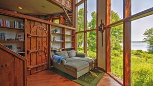 A cozy room with a large window, wooden bookshelves, a sofa, and a staircase. - 'dragonwood Castle' on Waterfront w/ Stunning View (Prospect Harbor)