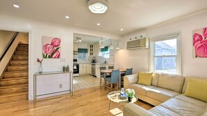 A modern living room with a sofa, coffee table, and artwork. A staircase leads to a kitchen area with a dining table and chairs.