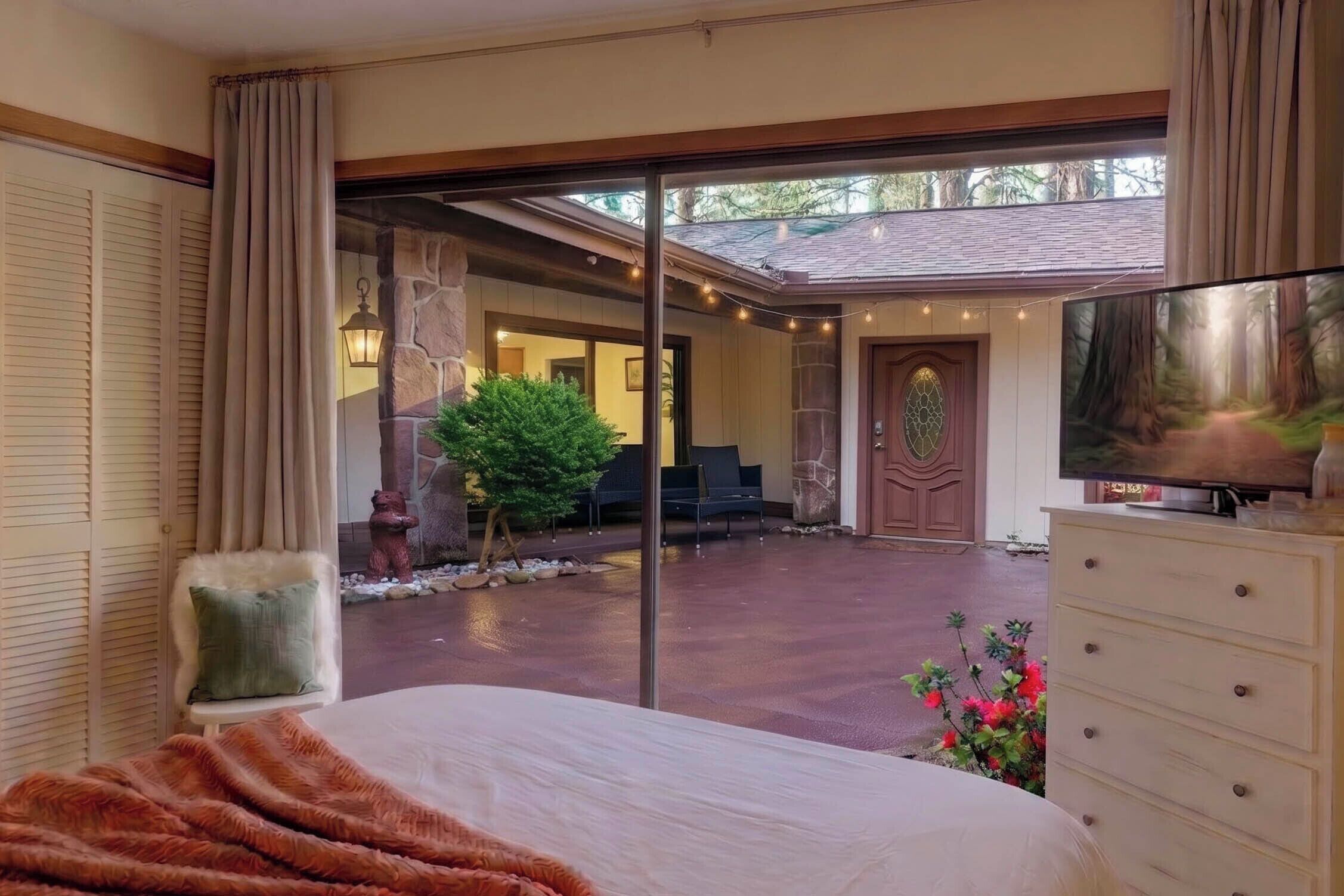 Un dormitorio con una cama grande, un aparador, un televisor y vistas a un patio exterior con un muro de piedra, una silla y un árbol pequeño.