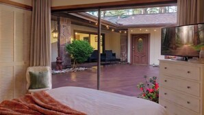 Un dormitorio con una cama grande, un aparador, un televisor y vistas a un patio exterior con un muro de piedra, una silla y un árbol pequeño.