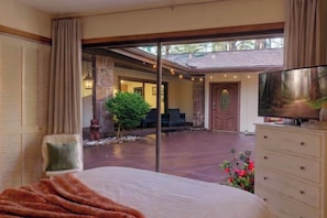 A bedroom with a large bed, a dresser, a television, and a view of an outdoor patio area with a stone wall, a chair, and a small tree. - Hot Tub + Fire Pit: Creekside Gatlinburg Getaway! (Gatlinburg)