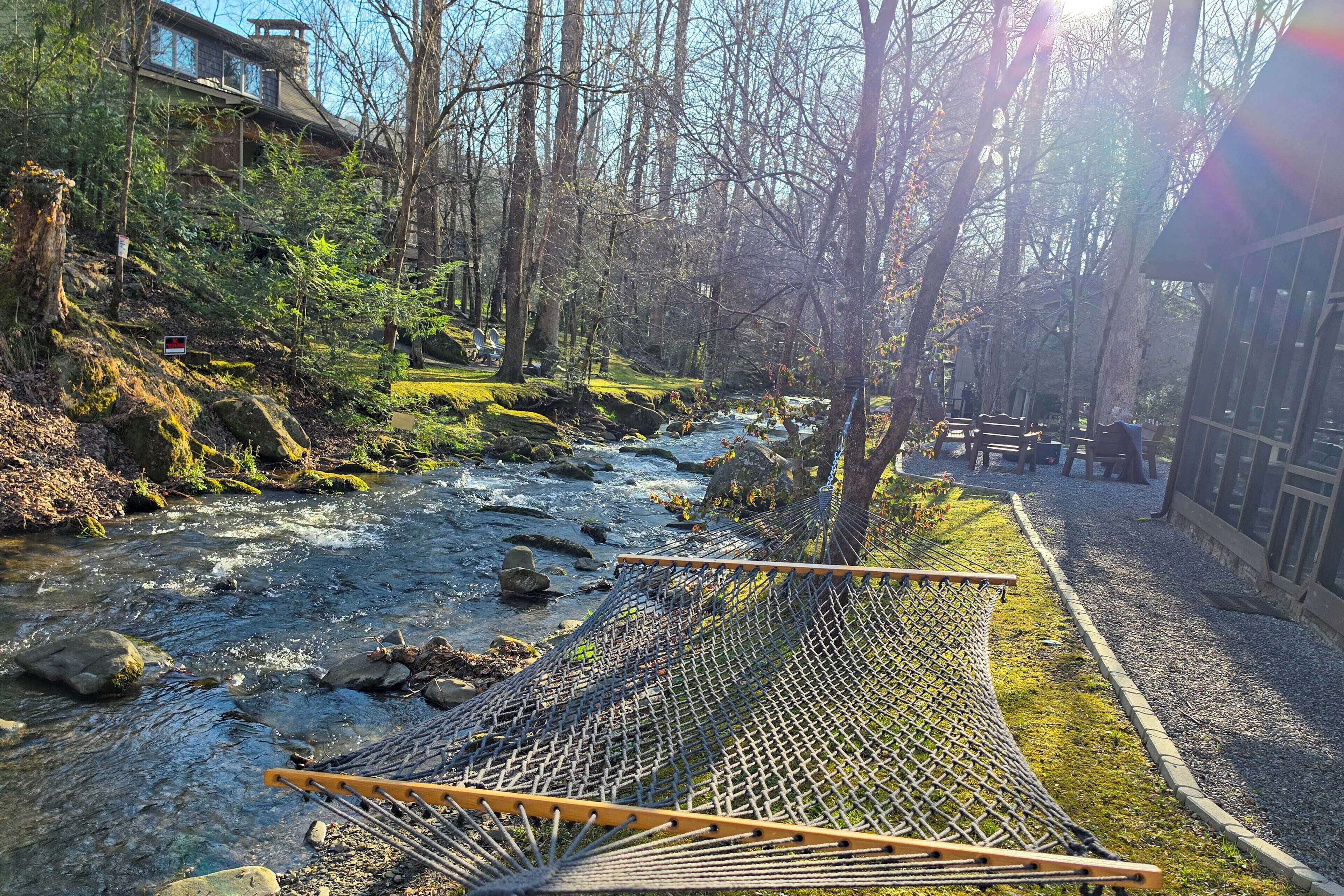 Een hangmat aan een rivier, omringd door bomen en een gebouw met grote ramen.