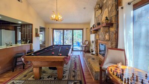 Una mesa de billar con un candelabro, una chimenea y un tablero de ajedrez en una sala con paredes de piedra y ventanas amplias.