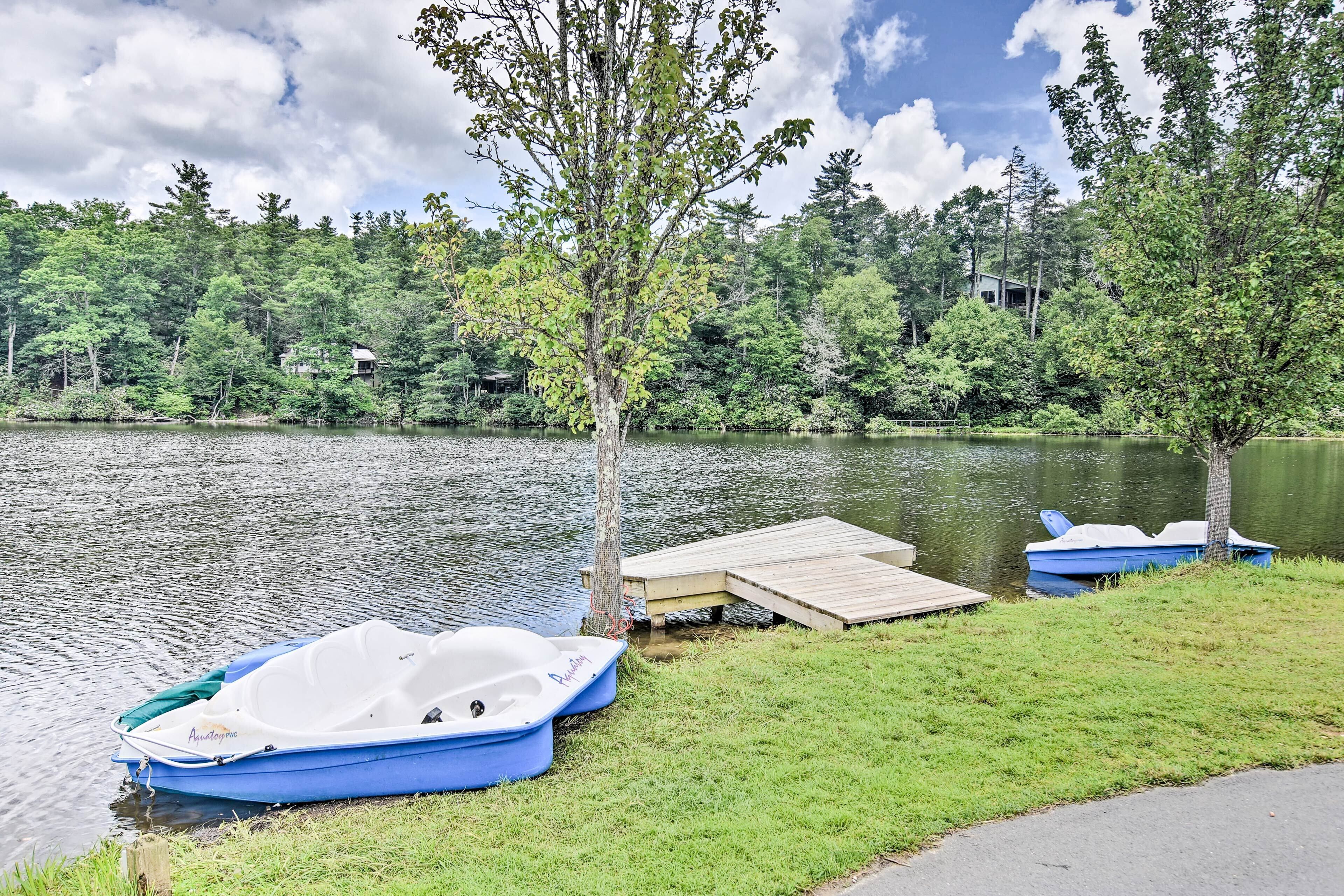 A lakeside scene with two boats docked at a wooden pier, surrounded by lush greenery and a forested backdrop.