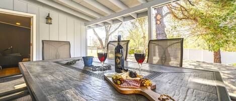 Un espacio al aire libre cubierto para comer, con una mesa dispuesta para dos personas, que incluye una tabla de madera con varios quesos y embutidos, dos copas de vino y una botella.