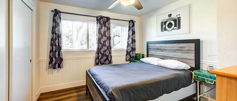 Une chambre à coucher comprenant un lit, un ventilateur de plafond, une fenêtre avec des rideaux, une caméra fixée au mur et une table de chevet.