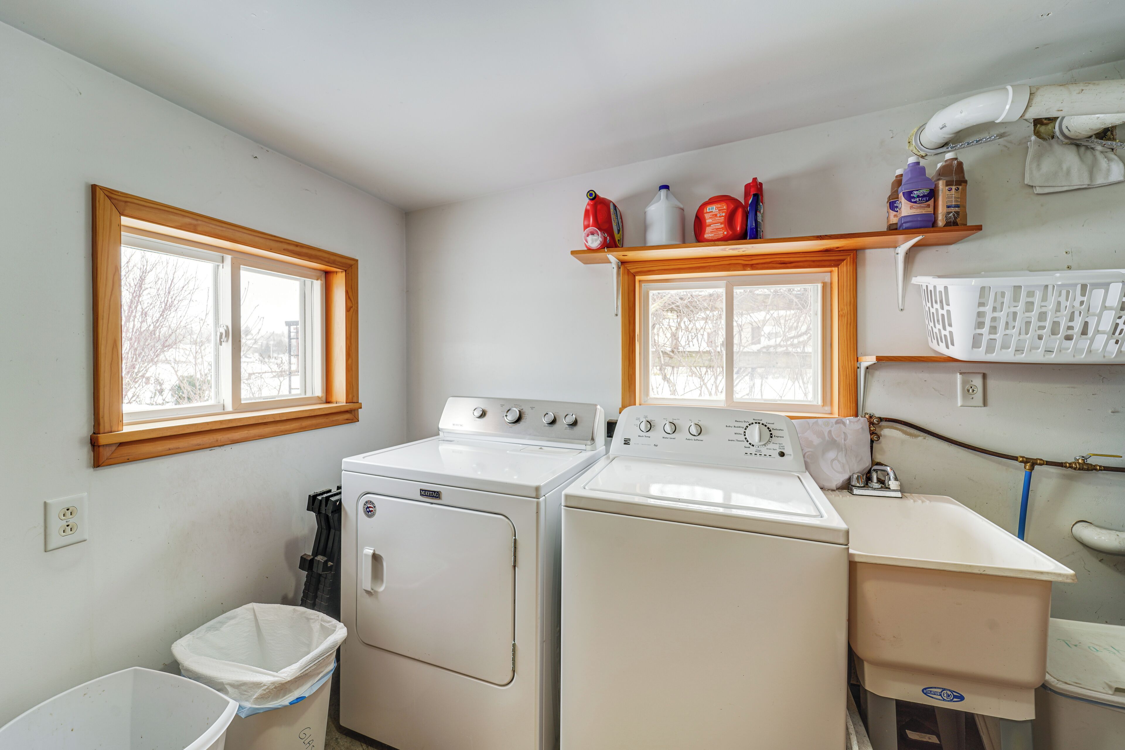 A laundry room with two white washers and dryers, a window, and a shelf with cleaning supplies.