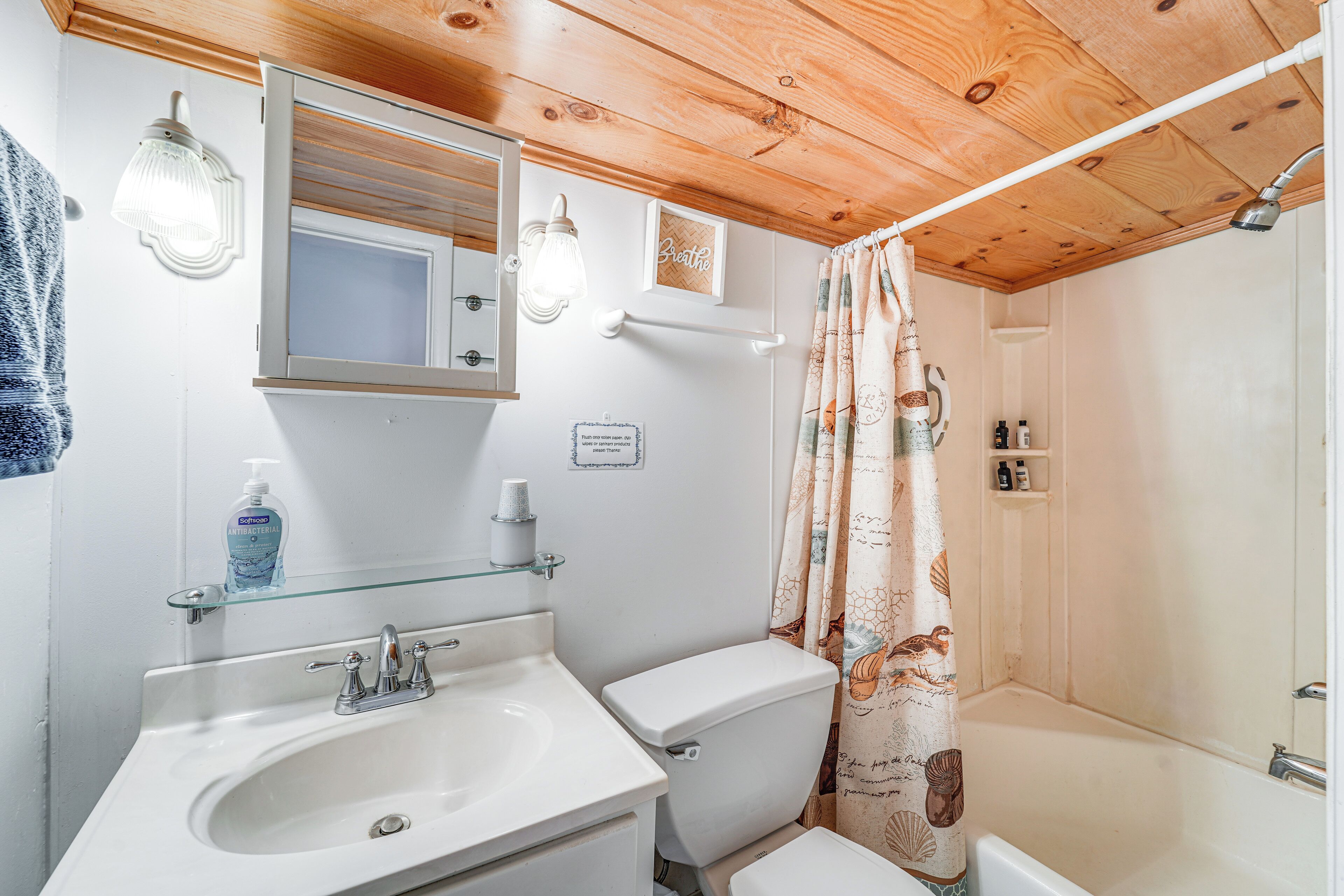 A bathroom with a wooden ceiling, a glass shelf above the sink, a toilet, and a shower curtain with a nautical theme.