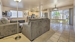 A living room with a gray sofa, a dining table with chairs, a kitchen area with cabinets and appliances, and a large window with blinds. - Beautiful St George Condo ~ 5 Mi to Downtown! (St. George)