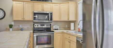 A modern kitchen with light wooden cabinets, stainless steel appliances including a refrigerator, oven, and microwave, and a clock on the wall.