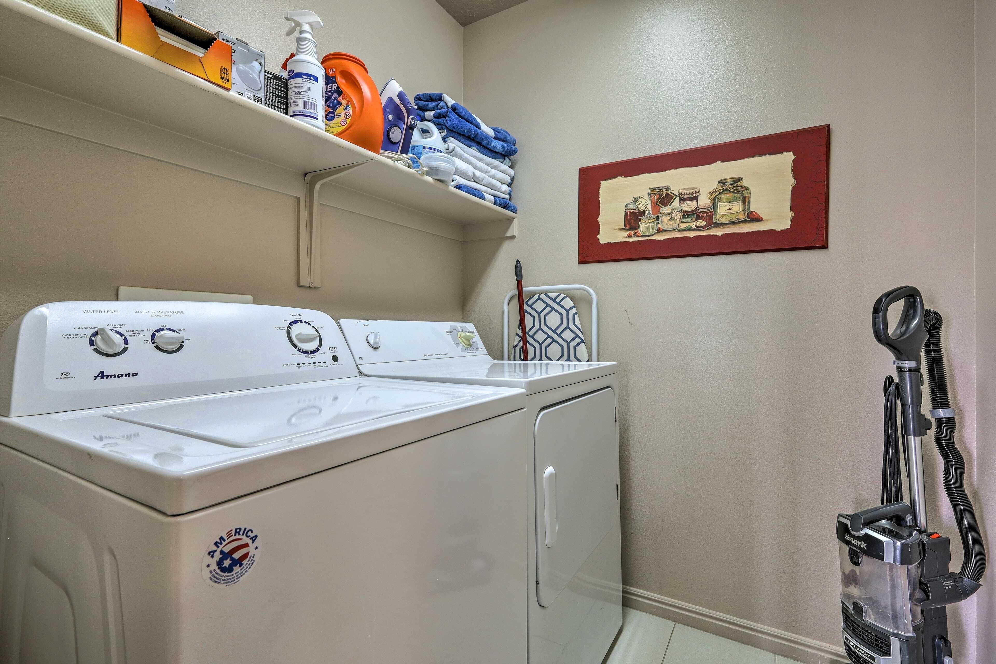 Un espace buanderie comprenant un lave-linge et un sèche-linge à chargement frontal, une étagère avec des produits de lessive, un aspirateur et un tableau encadré accroché au mur.