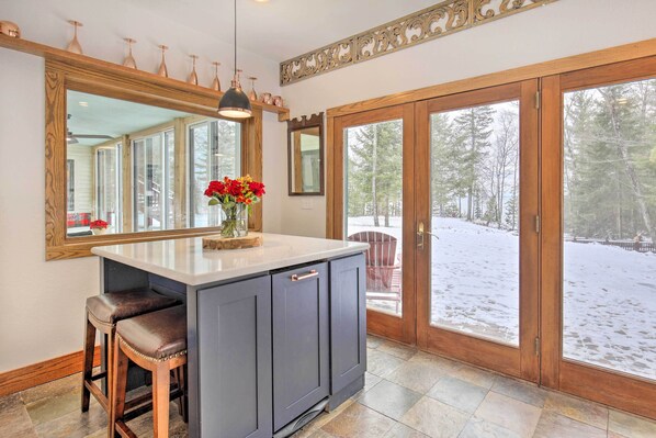 A kitchen with a large island, bar stools, a vase of red flowers, and a view of a snowy outdoor area through glass doors. - Cabin on Lake Superior ~ 11 Mi to Bayfield! (Washburn)