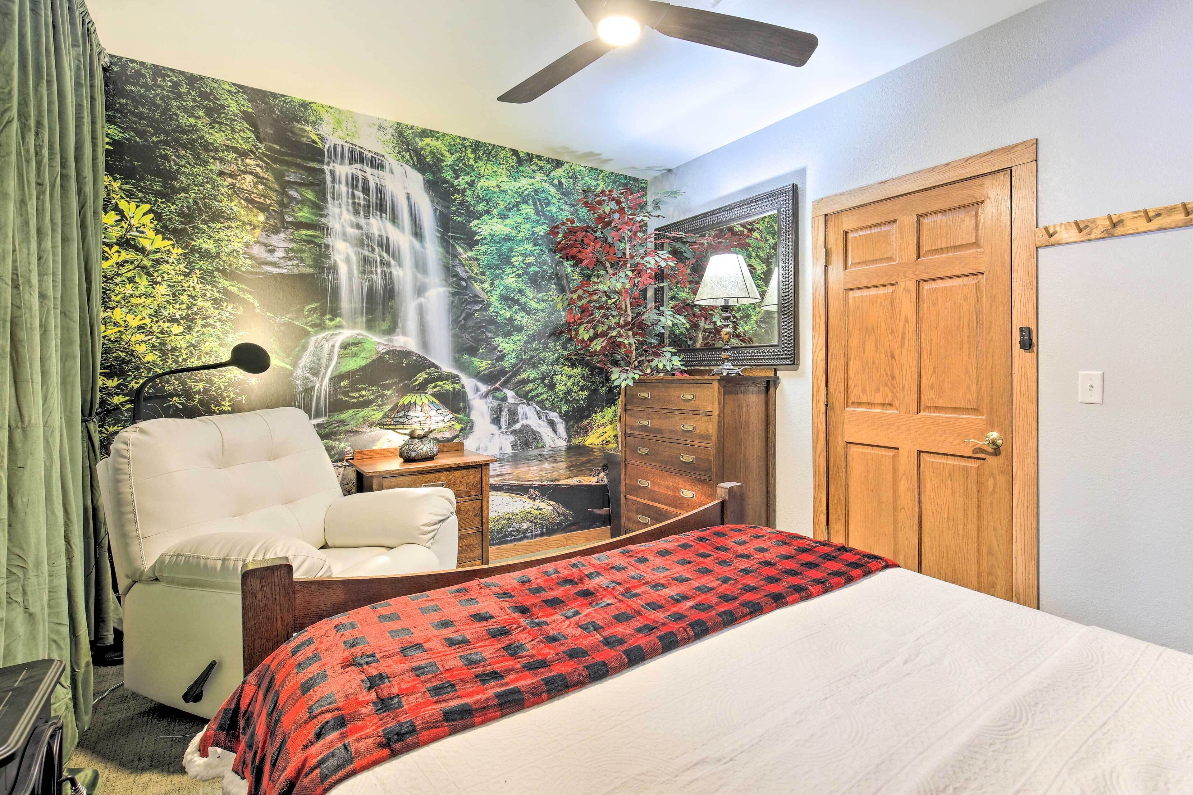 A bedroom with a bed, a chair, a desk, a mirror, a door, and a wall mural of a waterfall.