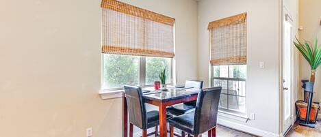 A dining area with a wooden table and chairs, two windows with blinds, and a potted plant.