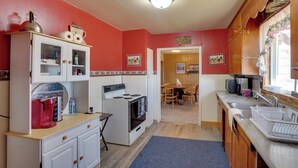 Een kleine keuken met een fornuis, spoelbak en kastjes. Door een open deur is een eethoek met een tafel en stoelen zichtbaar.