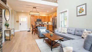 A spacious living room with a grey sofa, a wooden coffee table, and a dining area with wooden chairs and a chandelier. - Game Room & Wraparound Decks: Cullowhee Home! (Cullowhee)