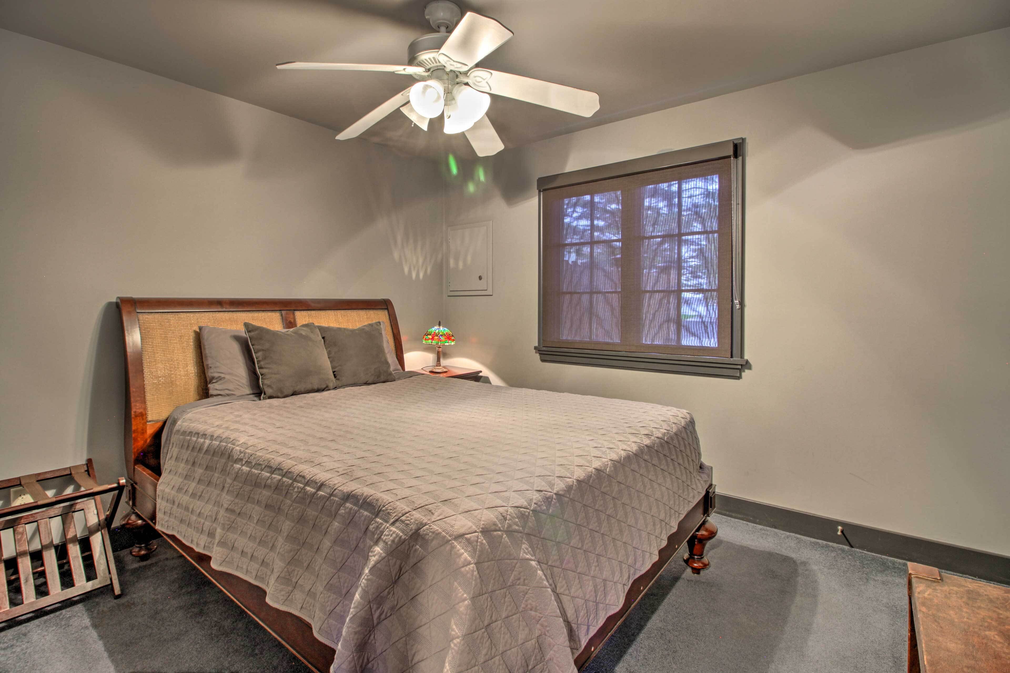 A bedroom with a bed, a nightstand, a crib, a ceiling fan, and a window with blinds.